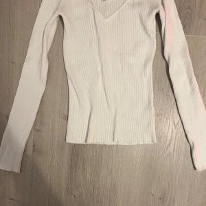 Vit ribbad långärmad topp - Snygg vit ribbad topp med v-ringning och lång ärm. Figurnära passform och stretchigt material som sitter bekvämt. Perfekt att styla till jeans eller kjol för en clean look.