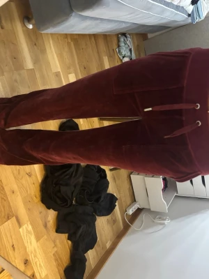 Vinröda mjukisbyxor fråm Juicy Couture  - Säljer ett par vinröda mjukisbyxor i mjuk velour med raka ben och dragsko i midjan. Byxorna har stora fickor fram och en bakficka med knappdetalj. Perfekta för chill dagar eller när du vill vara extra bekväm.