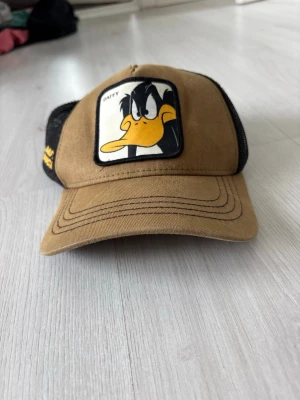 Brun truckerkeps med Daffy Duck - Säljer en brun och svart truckerkeps med mesh baktill och en stor patch framtill med Daffy Duck från Looney Tunes. Kepsen har böjd skärm och justerbar snapback. Materialet är en mix av bomull och polyester, perfekt för en avslappnad streetwear-look.