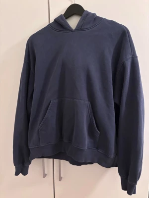 Mörkblå hoodie - En stilren mörkblå hoodie med huva och stor magficka framtill. Hoodien har ribbade muddar vid ärmslut och nederkant. Lite urtvättad 