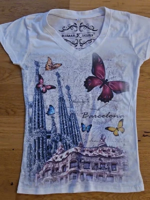 Vit t-shirt med Barcelona-tryck - Vit t-shirt från Ikebana K Design med färgglatt tryck med fjärilar🦋. T-shirten har v-ringning och korta ärmar. Materialet är mjukt och lätt. Använd ett fåtal gånger.