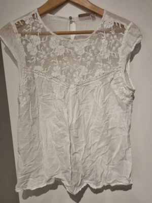 Vit spetsblus från KappAhl - Säljer en söt vit blus från KappAhl med transparent spets upptill och blommiga broderier. Blusen är ärmlös och har rund halsringning samt knapp i nacken. Perfekt för dig som gillar romantiska detaljer och en luftig känsla.
