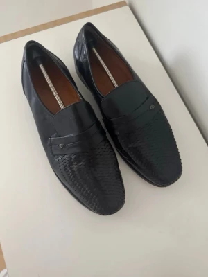 Svarta loafers i flätad läder - Snygga svarta loafers i läder med flätad detalj på tån och klassisk penny strap. Skorna har en stilren och tidlös design med låg klack och rundad tå. Perfekta för dig som vill ha en clean look med lite edge.