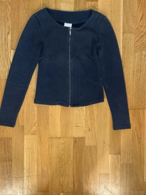 Mörkblå strukturerad cardigan - Mörkblå cardiganjacka med lång ärm och rund halsringning. Jackan har en diskret struktur i tyget och stängs med dragkedja framtill. Passformen är rak och enkel, vilket gör den lätt att matcha med olika outfits.