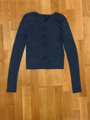Mörkblå ribbad kofta med knappar - Snygg mörkblå ribbad kofta med långa ärmar och rund halsringning. Koftan har en croppad passform och knäpps med svarta knappar framtill. Perfekt att styla över ett linne eller t-shirt för en chill look.