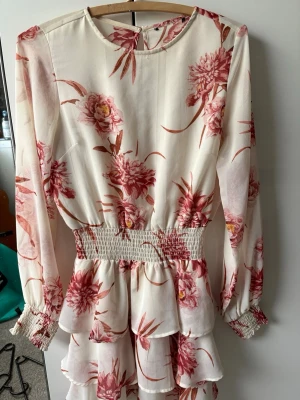 Blommig kortklänning med volanger - Söt kortklänning i vitt med rosa blommönster och långa ärmar. Har använd ett antal gånger. Har svårt att släppa denna klänningen men tänker att det är dags då den är för kort där bak på mig. Denna säljs ej längre då priset kan va högt😔