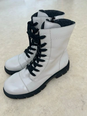 Vita kängor med svarta snören - Snygga vita kängor i glansigt material med svarta snören och grov sula. Kängorna har dragkedja på sidan och svart innerfoder. 