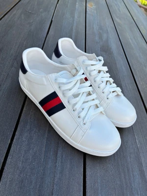 Gucci Ace vita - Snygga stilrena vita Gucci Ace skor med röda och blåa detaljer. skorna har blå baksida på båda sidor samnt blåa och röda ränder vid sidan vilket ger dem en extremt stilren look.