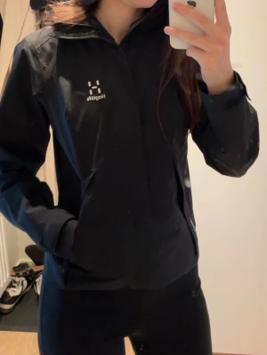 Svart vindjacka från Haglöfs - Snygg svart vindjacka från Haglöfs med minimalistisk design och diskret logga på bröstet. Jackan har dragkedja framtill, två sidofickor och är tillverkad i ett lätt och smidigt material. Perfekt för blåsiga dagar och outdoor-aktiviteter.