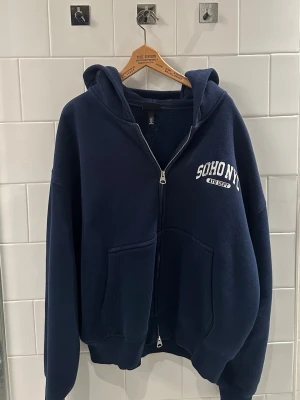 Mörkblå SOHO NYC hoodie - Mörkblå hoodie med dragkedja och vit text 'SOHO NYC ATH DEPT' på bröstet. Hoodien har huva och två stora fickor framtill. Perfekt för en avslappnad streetstyle-look. Materialet är mjukt och bekvämt, troligen bomullsblandning.