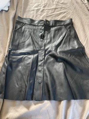 Svart skinnkjol med knappar - Snygg svart kjol i skinnimitation med knappar framtill och hög midja. Kjolen har fickor på sidorna och en lätt utställd form som ger en cool och trendig look. Perfekt att matcha med en stickad tröja eller t-shirt.