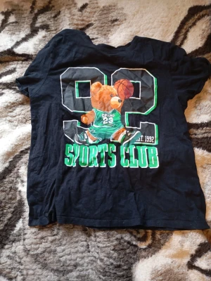 Svart t-shirt med björn och tryck - Cool svart t-shirt med stort tryck på framsidan av en björn i basketkläder, texten 'SPORTS CLUB' och siffran 92. T-shirten har rund hals och korta ärmar. Perfekt för dig som gillar sportiga och lekfulla motiv. Knappt använd. Pris kan diskuteras🙌. Mvh Rasmus.