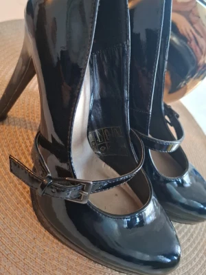 Svarta lackpumps med rem från Duffy - Snygga svarta pumps i lack från Duffy med klassisk Mary Jane-rem och rundad tå. Skorna har en elegant klack och justerbart spänne vid sidan. Perfekta för dig som vill ha en stilren och trendig look.