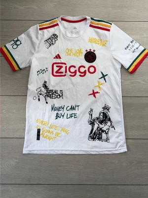 Vit t-shirt med Bob Marley-tryck - Unik vit t-shirt med färgglada detaljer på ärmarna och flera tryck inspirerade av Bob Marley och reggae. Framsidan har Ziggo-logga, Ajax-märke och citat, medan baksidan pryds av ett stort Bob Marley-porträtt och texten 'Live the life you love'. 