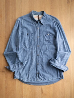 Nudie Jeans Workers Shirt, size M - Säljer en klassisk blå jeansskjorta med två bröstfickor och knappar framtill. Skjortan har lång ärm och en avslappnad passform. Perfekt till ett par svarta jeans eller över en t-shirt för en chill look.