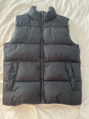 Svart Tommy Hilfiger dunväst  - Snygg svart dunväst med hög krage och quiltad design. Västen har två fickor framtill och stängs med dragkedja. Liten diskret logga på bröstet. Perfekt för lager-på-lager och passar till många olika stilar. Storlek 164/XS