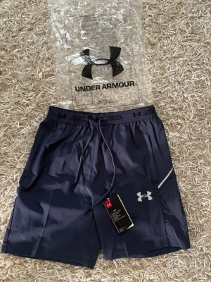 Blå träningsshorts från Under Armour - Snygga marinblå träningsshorts från Under Armour med snörning i midjan och logga på vänster ben. Shortsen har meshpanel på sidan för extra ventilation och en sportig look. Perfekta för gymmet eller löprundan.