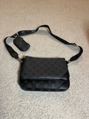 Louis Vuitton svart axelväska monogram - Snygg svart axelväska från Louis Vuitton med klassiskt monogrammönster i grått och svart. Väskan har justerbar axelrem och en liten avtagbar ficka. Tillverkad i läder med silverfärgade detaljer. Perfekt för dig som vill ha en stilren och lyxig accessoar.