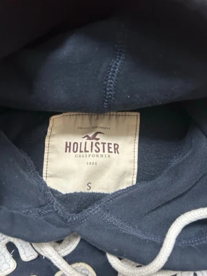 Mörkblå hoodie från Hollister - Snygg mörkblå hoodie från Hollister med broderad logga och texten 'Cali Dreams' på bröstet. Hoodien har en klassisk huva med vita dragsnören och är tillverkad i mjukt bomullsmaterial. Perfekt för en avslappnad och trendig look.
