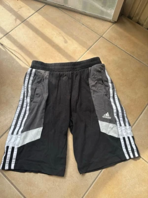 Svarta Adidas shorts med vita ränder - Säljer ett par svarta Adidas shorts med klassiska vita ränder längs sidorna och gråa detaljer. Shortsen har elastisk midja och två fickor framtill. Perfekta för träning eller chill dagar. Adidas-logga på vänster ben.