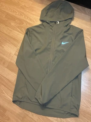 Nike windbreaker - Nike Dri-FIT vindjacka i olivgrönt med huva och hel dragkedja framtill. Jackan har en diskret Nike-logga på bröstet och är tillverkad i ett lätt, vattenavvisande material som andas. Perfekt för blåsiga dagar och sportiga outfits.