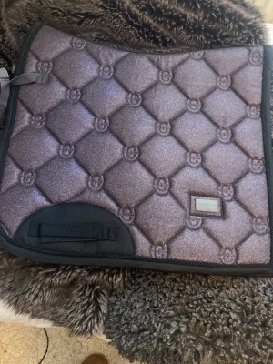 Glittrigt schabrak i lila och svart - Säljer ett schabrak med glittrig lila yta och svart kant. Schabraket har quiltat mönster med små emblem i rutorna och en svart förstärkning vid sadelgjorden. Perfekt för dig som vill sticka ut i stallet.