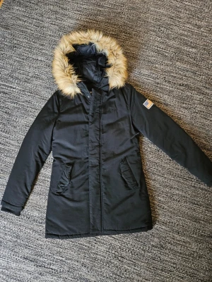 SVEA Miss Lee Jacket - Snygg och varm vinterjacka från SVEA, modell Miss Lee Jacket, i klassisk svart färg. Jackan har en fiqursydd passform, avtagbar huva med fyllig fuskpäls, dragkedia med knappslå och två framfickor samt två innerfickor. Broderad SVEA-logga på pröstet samt den ikoniska USA-patchen på ärmen. Mvcket fint skick!
