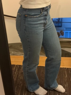 Blå raka jeans från Levi's - Säljer ett par klassiska blå jeans från Levi's med rak passform och hög midja. Jeansen har fem fickor, knappgylf och är tillverkade i ett slitstarkt denimtyg. Perfekta för en avslappnad och tidlös stil.
