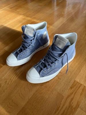 Converse Chuck 70 Stars - Blå höga sneakers - Converse Chuck 70 Stars - Blå höga sneakers. Snygga höga sneakers i blå canvas med vita gummisulor och tåhättor. Skorna har klassisk snörning och rundad tå. Perfekta för dig som gillar en retro och avslappnad stil. Passar till jeans eller shorts och ger en cool vibe till outfiten.