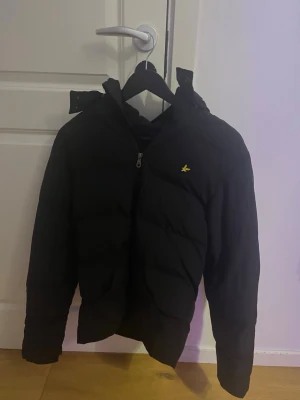 Svart pufferjacka med gul logga - Snygg svart pufferjacka med dragkedja och huva. Jackan har en gul broderad logga på bröstet och quiltad design som ger en modern look. Perfekt för kalla dagar när du vill hålla värmen och ändå se stilren ut.