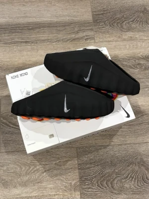  Mind svarta tofflor - Säljer ett par svarta Mind tofflor med en futuristisk design och synlig silverfärgad Swoosh på sidan. Sulan har färgglada detaljer undertill som ger en cool kontrast. Perfekta för dig som gillar streetwear och unika skor. Har Andvänd 3-5 gånger Och underifrån är det lite brunt och små repor därför kan priset diskuteras 