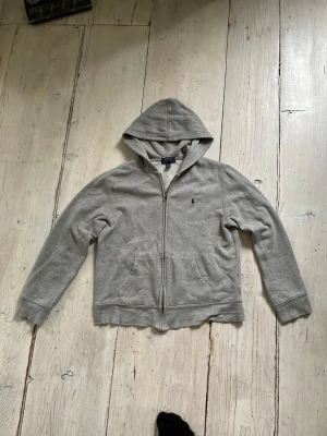 Grå hoodie från Polo Ralph Lauren - Klassisk grå hoodie från Polo Ralph Lauren med dragkedja framtill och två fickor. Mjuk insida och broderad logga på bröstet. Perfekt för en avslappnad och stilren look. Storlek L (14-16).