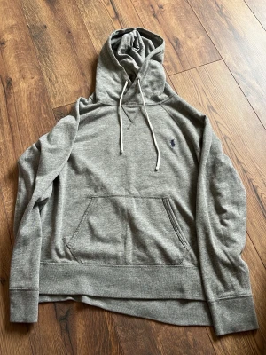 Grå hoodie med huva och magficka - Säljer en grå hoodie med huva och vita dragsnören. Hoodien har en klassisk magficka och en liten broderad logga på bröstet. Tillverkad i mjukt sweatshirtmaterial, perfekt för en avslappnad stil.