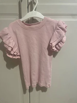 Ljusrosa topp med volangärmar - Supersöt ljusrosa topp med korta volangärmar. Toppen har rund halsringning och är gjord i ett mjukt bomullsmaterial. Perfekt för dig som vill ha en gullig och trendig look med extra detaljer på ärmarna.