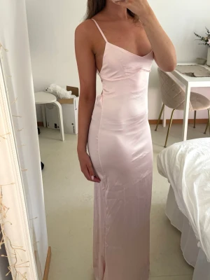Ljusrosa balklänning med smala band - Elegant ljusrosa långklänning i satinliknande material med smala axelband och v-ringad urringning. Klänningen har en figurnära passform och slät yta som ger en stilren look. Det tunna och stretchiga materialet formar sig fint efter kroppen och ger en smickrande silhuett. Perfekt för dig som vill ha en snygg klänning till bal, gala eller bröllop.