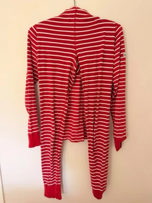 Polarn o pyret pyjamas  - Mysig onesie pyjamas i rött med vita horisontella ränder. Långärmad modell med rund halsringning och muddar vid ärmar och benslut. Tillverkad i mjukt, stretchigt material som passar perfekt för slappa dagar hemma.