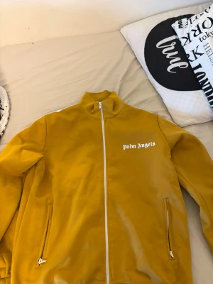 Gult tracksuit-set från Palm Angels - Snyggt gult tracksuit-set från Palm Angels med vit dragkedja och vita detaljer längs sidorna. Jackan har hög krage och två fickor, byxorna har resår i midjan och sidofickor. Båda delarna har Palm Angels-logga tryckt i vitt.