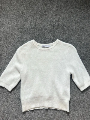 Vit stickad topp från Zara - Snygg vit stickad topp från Zara med rund halsringning och halvlånga ärmar. Tröjan har ribbade muddar vid hals, ärmslut och nederkant. Perfekt att styla med jeans eller kjol för en clean look.