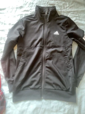 Svart träningsjacka från Adidas - Snygg svart träningsjacka från Adidas med dragkedja framtill och hög krage. Jackan har två sidofickor och den klassiska Adidas-loggan på bröstet. Perfekt för träning eller chill. Tillverkad i ett mjukt och stretchigt material.
