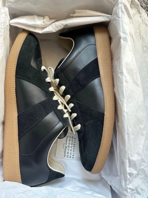 Svarta sneakers med mockadetaljer - Snygga svarta sneakers med ovandel i läder och mocka, vita snören och klassisk gummisula i beige. Skorna har en stilren design med svarta paneler och låg profil. Perfekta för dig som gillar minimalistisk och tidlös stil.