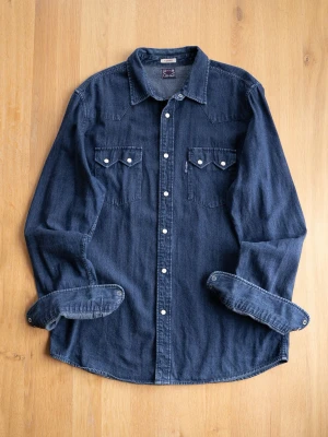 Navy Western Shirt, size XL - Säljer en klassisk mörkblå jeansskjorta med vita knappar och två bröstfickor med lock. Skjortan har lång ärm och en avslappnad passform. Perfekt till ett par svarta jeans eller över en t-shirt.