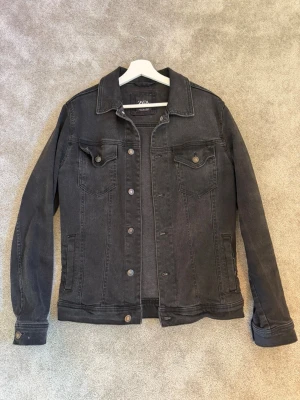Svart jeansjacka från Zara - Snygg svart jeansjacka från Zara med klassisk krage, knäppning framtill och två bröstfickor med lock. Jackan har raka ärmar och diskreta sömmar som ger en stilren look. Perfekt att slänga på sig över en hoodie eller t-shirt. Säljes då den inte passar längre. 