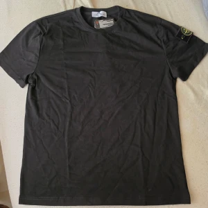 Svart t-shirt från Stone Island - Säljer en svart t-shirt från Stone Island med den klassiska loggan på ärmen. T-shirten har rund hals och korta ärmar. Tillverkad i mjuk bomull för en skön och avslappnad stil. Perfekt till jeans eller shorts.