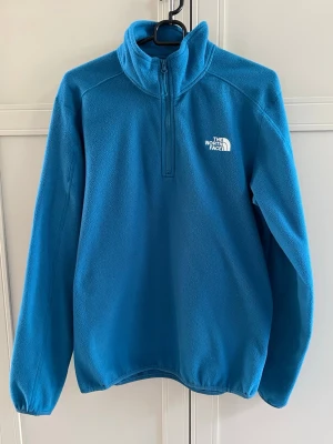 Blå fleece från The North Face - Blå fleece med halv dragkedja från The North Face. Tröjan har hög krage, lång ärm och logga broderad i vitt på bröstet och ryggen. Tillverkad i mjukt fleecematerial som håller dig varm under kyliga dagar.