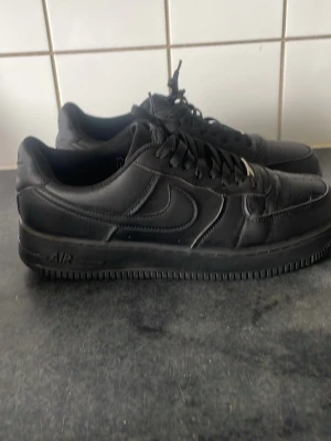 Svarta Nike Air Force 1 sneakers - Klassiska helsvarta Nike Air Force 1 sneakers i läder med låg profil och tydlig swoosh på sidan. Skorna har svart sula, svarta skosnören och diskret AIR-detalj på sulan. Perfekta för en clean och stilren look.