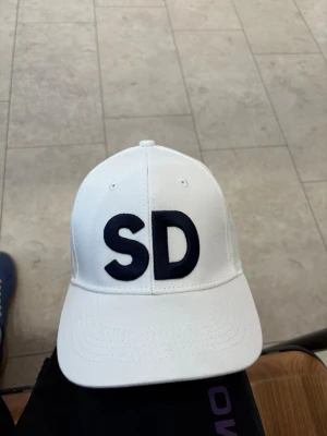 Sverigedemokraterna keps  - Två kepsar i klassisk modell, en vit med mörkblå broderade bokstäver 'SD' och en marinblå med ljusblå broderade bokstäver 'SD'. Båda har böjd skärm och justerbar baksida. Materialet ser ut att vara bomull eller polyester.