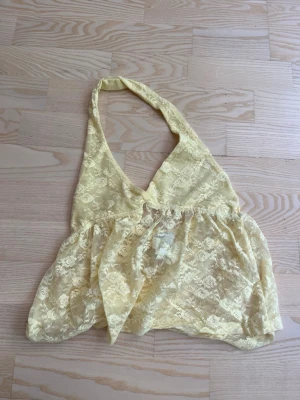 Gul spetsig halterneck topp - Säljer en ljusgul halterneck topp i spets med v-ringning och knyt i nacken. Toppens material är genomskinligt och har ett blommigt mönster. Perfekt för dig som gillar unika och luftiga plagg.