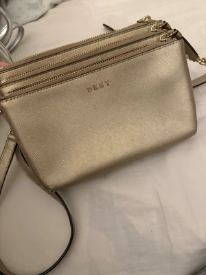 Guldig axelväska från DKNY - Snygg guldig axelväska från DKNY med tre dragkedjefack och justerbar axelrem. Väskan har en stilren design i glansigt material och logga framtill. Perfekt för att bära det viktigaste och ger en lyxig touch till din outfit.