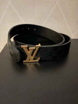 Elegant svart designbälte med guldspänne - Elegant designerbälte med ett karaktäristiskt rutmönster och ett utmärkande guldspänne. Bältet är tillverkat av högkvalitativt läder och förenar hållbarhet med stil. Längden är angiven som storlek 44/110. Perfekt för att lyfta vilken outfit som helst.