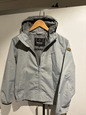 Ljusgrå regnjacka från Barbour - Ljusgrå regnjacka från Barbour med huva och dragkedja framtill. Jackan har elastiska muddar, två sidofickor och meshfoder. Vattentät och med diskreta gula detaljer på huva och ärm. Perfekt för regniga dagar.
